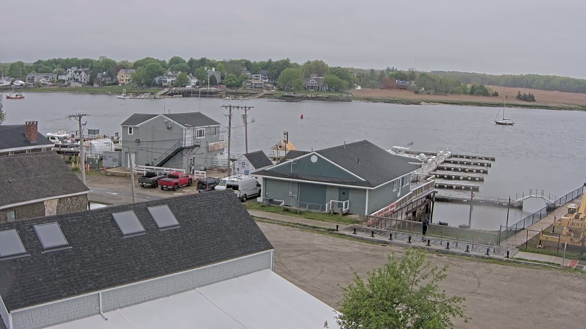 An overcast day shows a waterfront scene with buildings, docked boats, and a sailboat on the river, all under a gray sky.