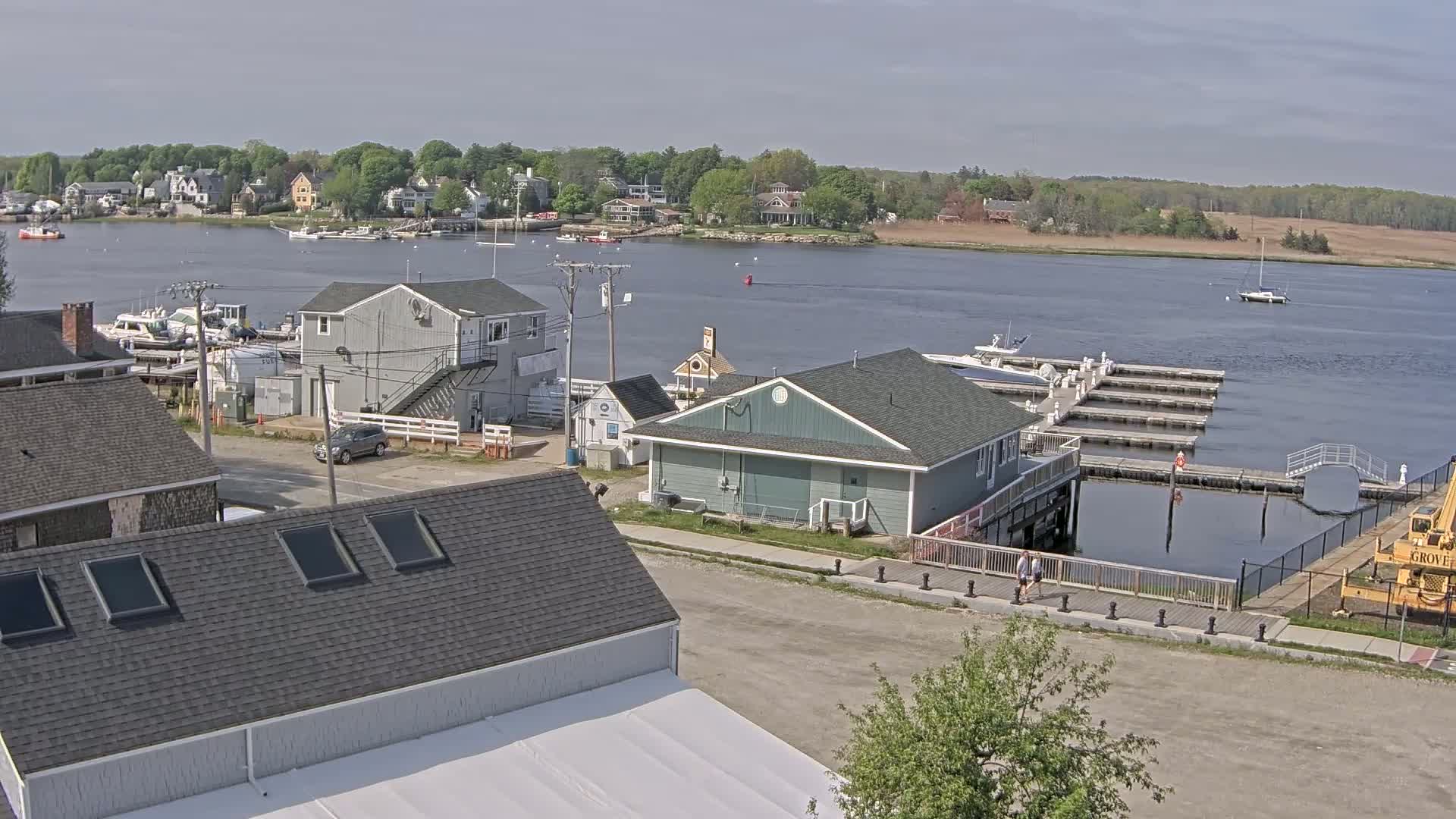 A waterfront town on a partly sunny day shows several buildings near a marina with boats docked, and a person walking along a walkway.