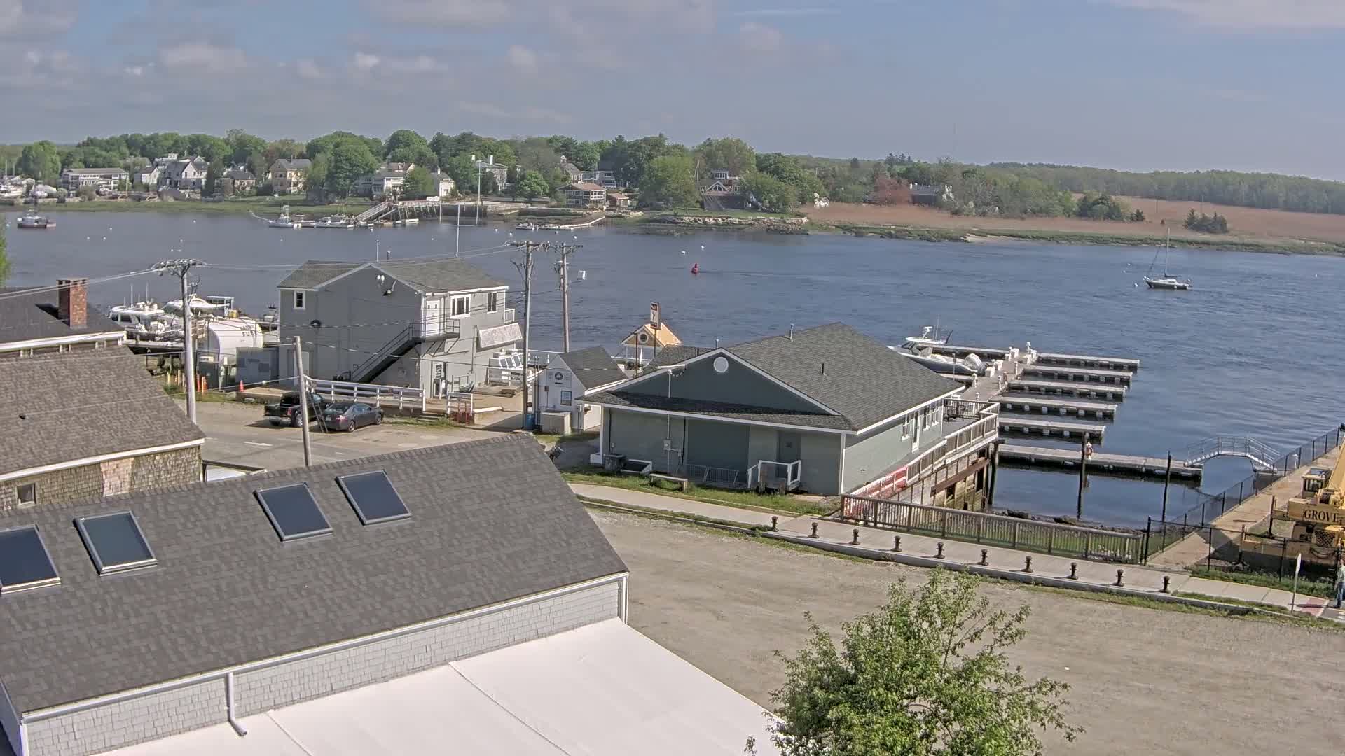 A waterfront view shows a small town with gray buildings near a calm river, boats docked at piers, and a partly cloudy sky.