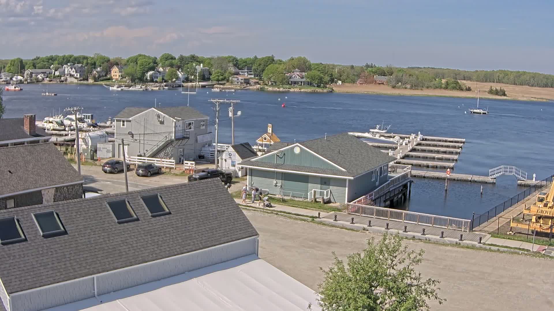 A waterfront town on a sunny day with boats docked at a marina, houses, and people walking along a paved walkway.