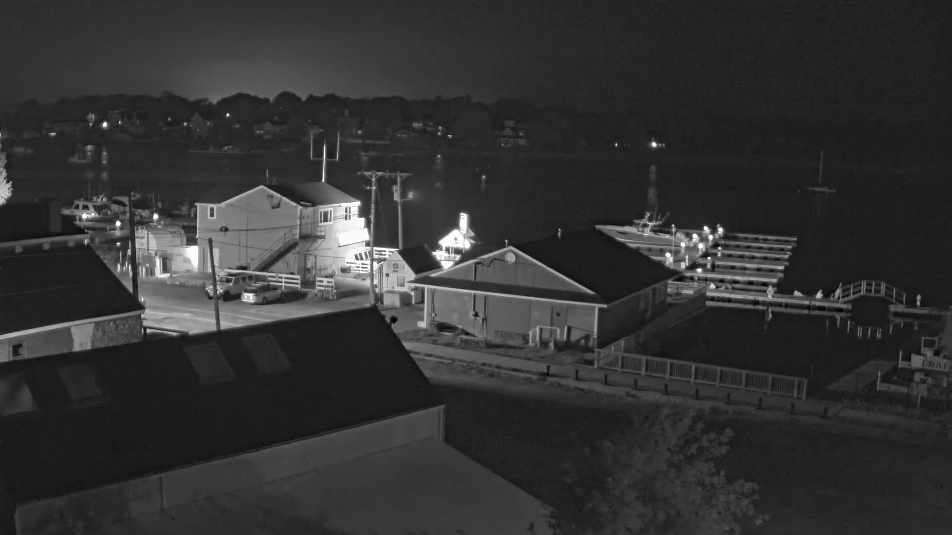 A nighttime, black-and-white view of a waterfront area with buildings, docks, boats, and cars, under a clear, dark sky.
