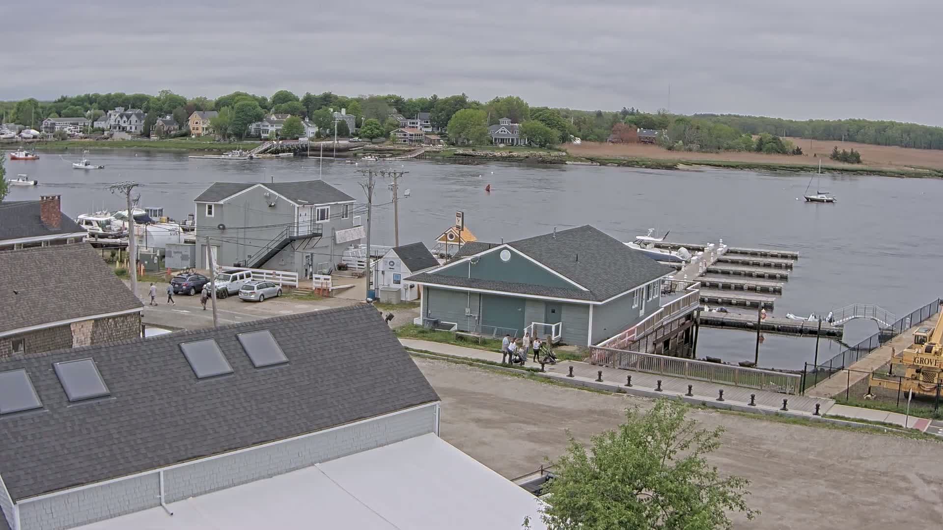 A waterfront town on an overcast day features several buildings, a marina with boats docked, and people walking near a roadway.