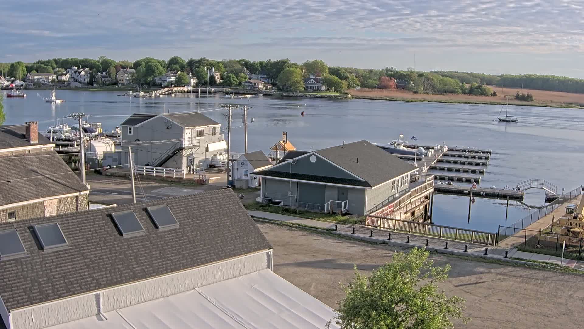 Under a partly cloudy sky, a waterfront town with several buildings, a marina filled with boats, and a calm river is viewed from an elevated perspective.