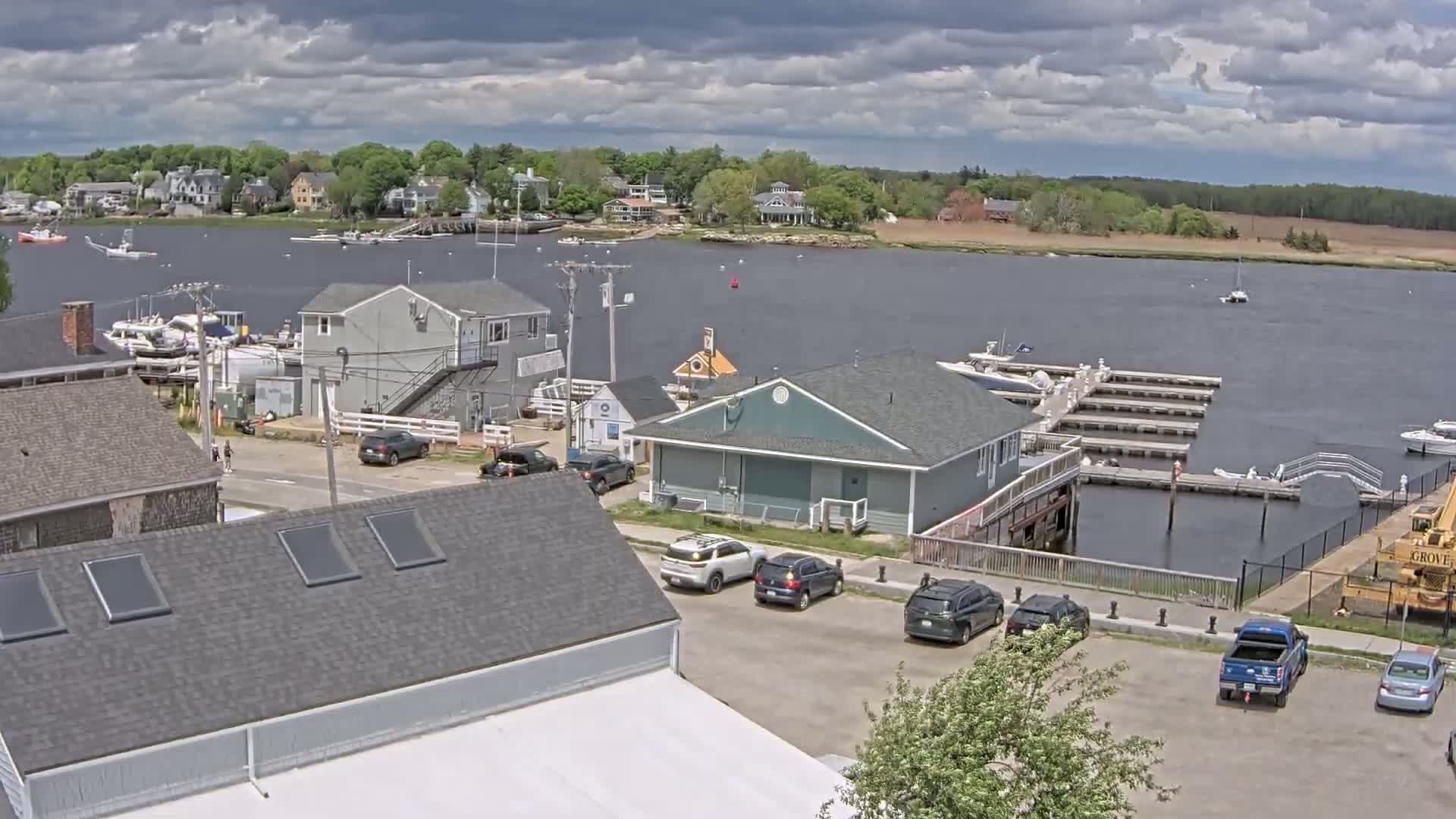 A partly cloudy day reveals a waterfront scene with houses, boats docked at a marina, and cars parked in a lot next to a building.