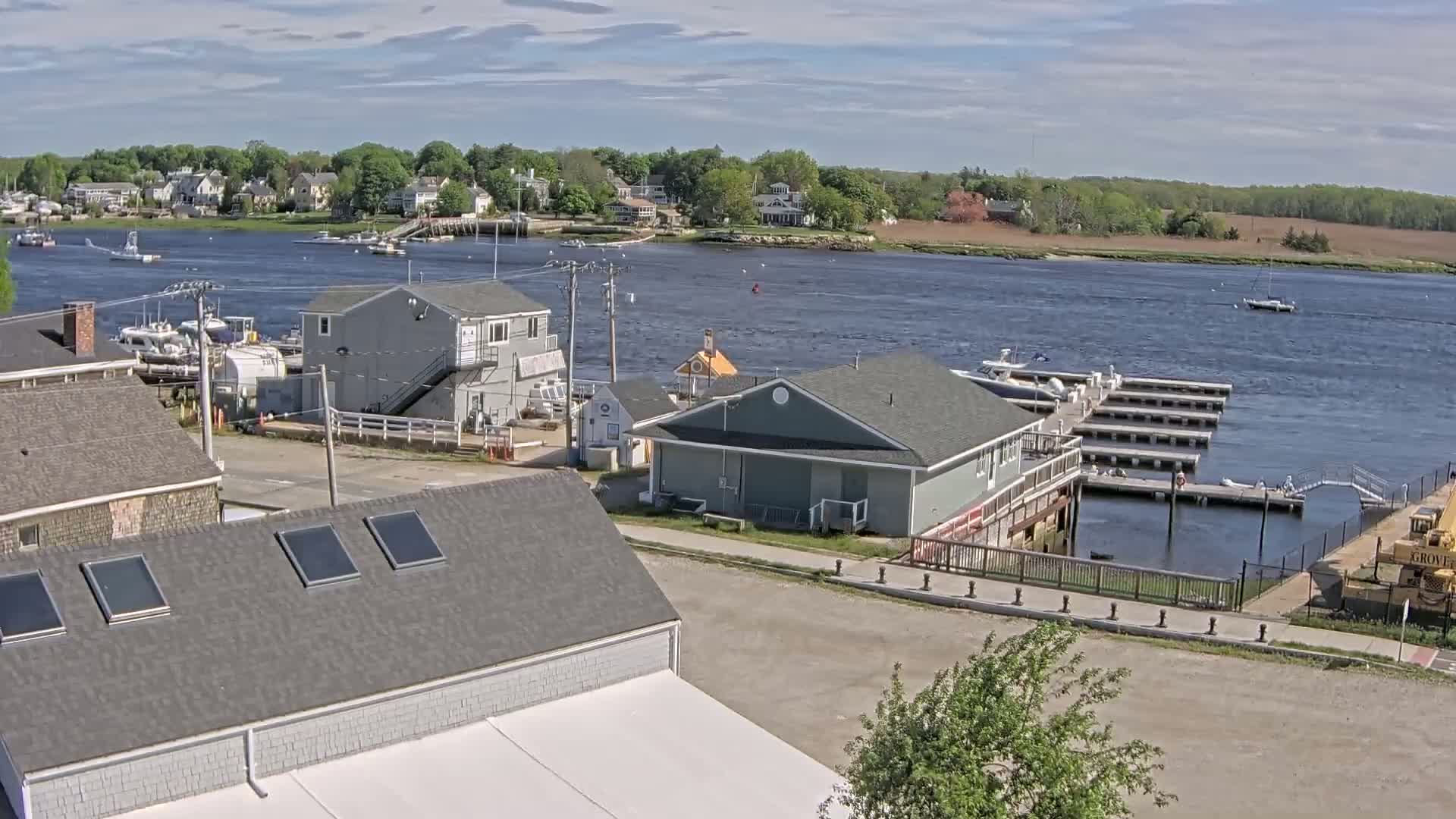 A waterfront scene on a partly cloudy day shows a small town with several buildings, boats docked at a marina, and a body of water extending to a treeline in the distance.