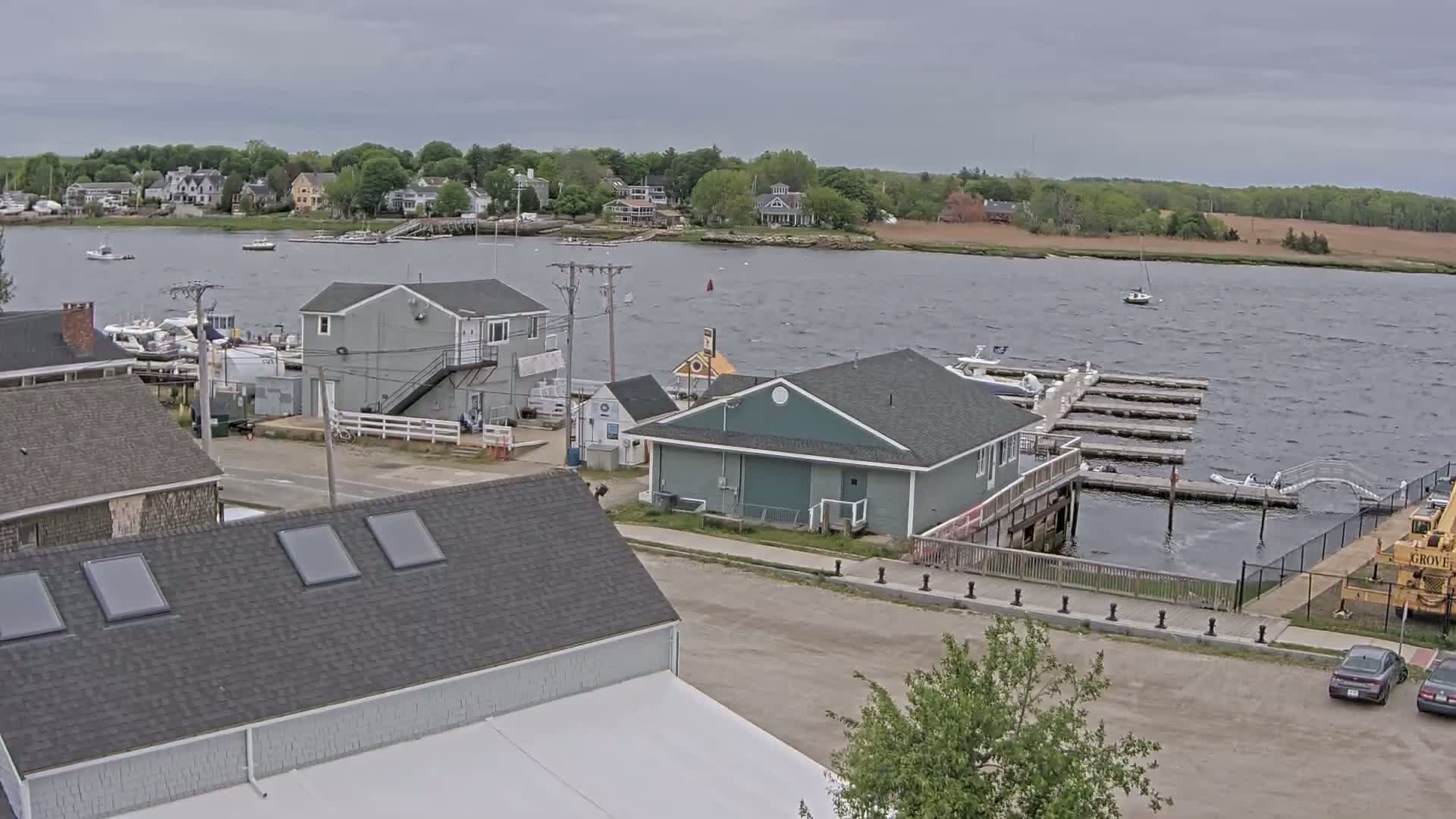 A waterfront scene on a cloudy day shows a marina with boats and docks, several buildings, and a parking lot, with a body of water in the background.