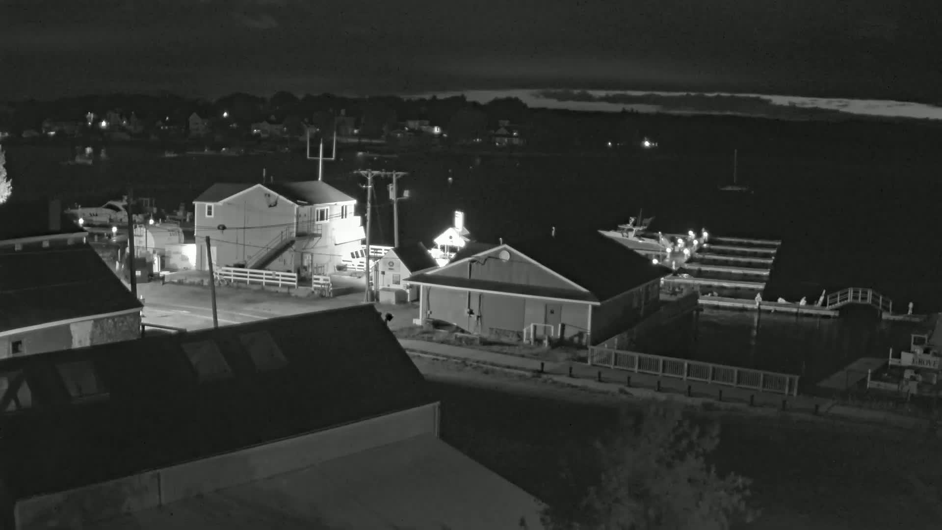 A nighttime view of a waterfront community with several buildings near a harbor containing boats at docks under a partly cloudy sky.