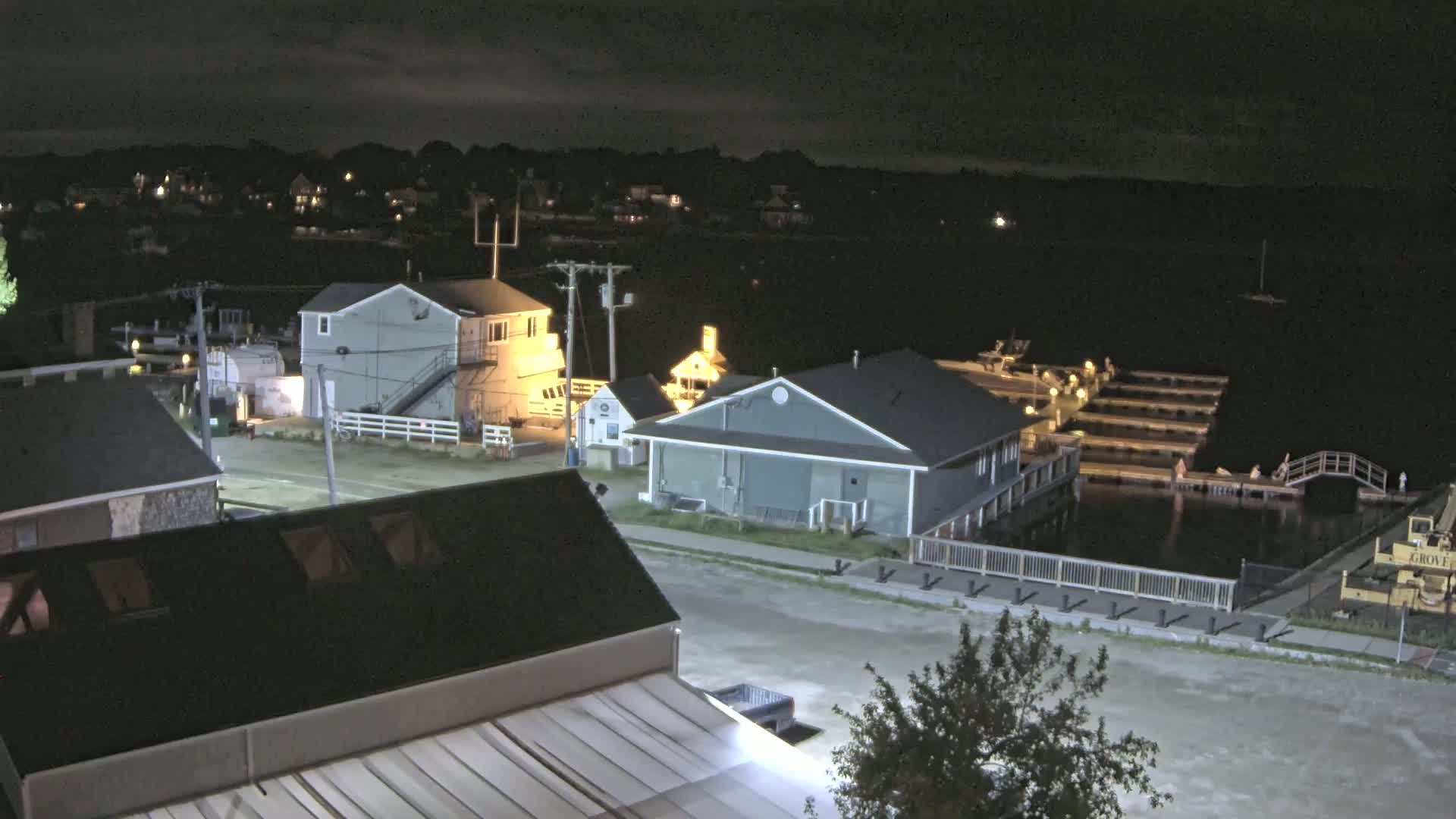 An elevated nighttime view shows a waterfront area with several buildings, docks, and boats under a partly cloudy sky.
