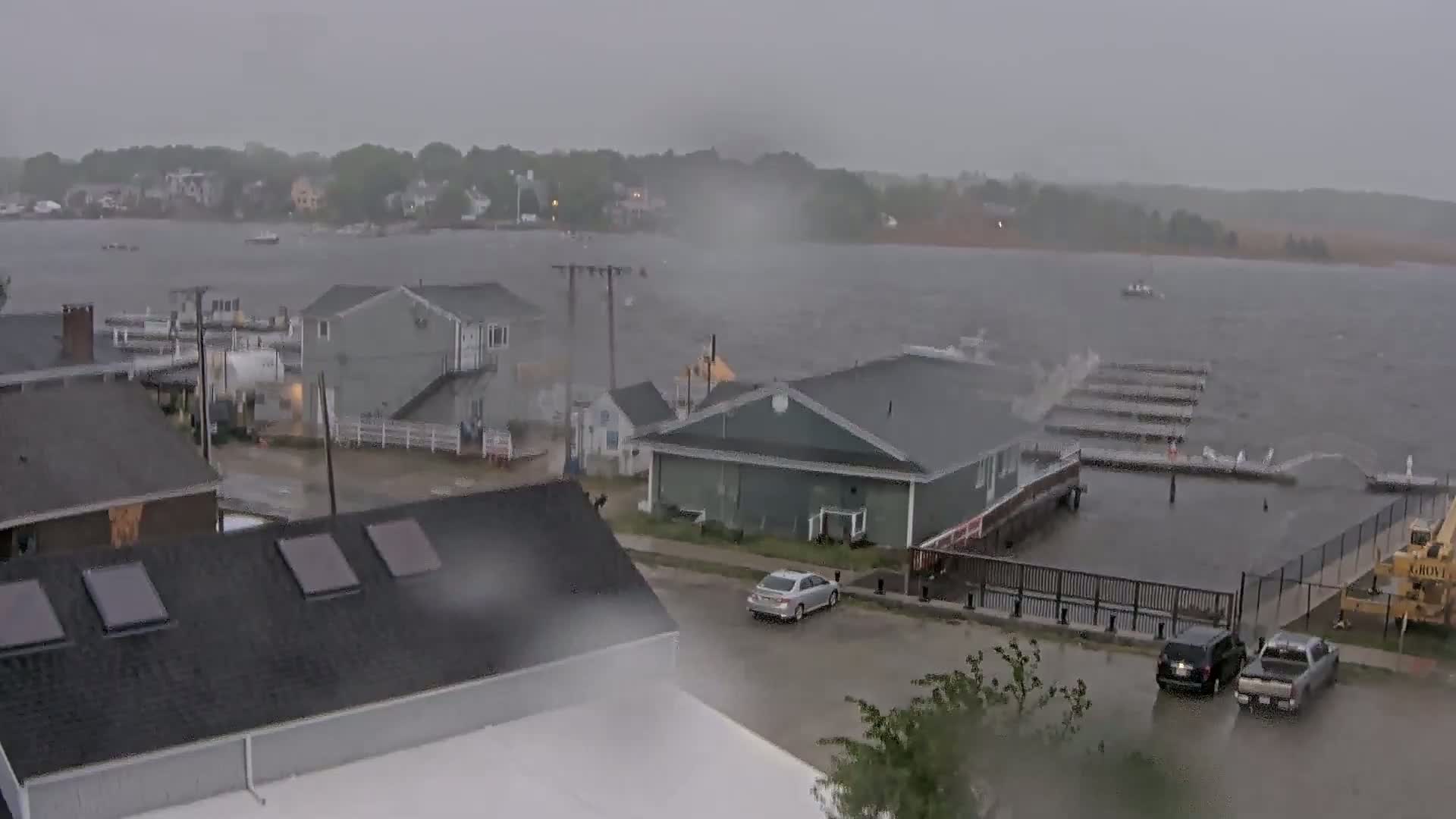 A waterfront area with houses and a marina is experiencing heavy rain and high water levels, with waves crashing against the docks.