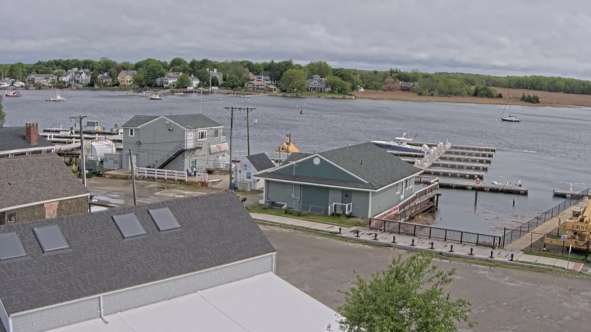 A waterfront view on an overcast day shows a small town with houses, a marina with boats docked, and several buildings near the water.