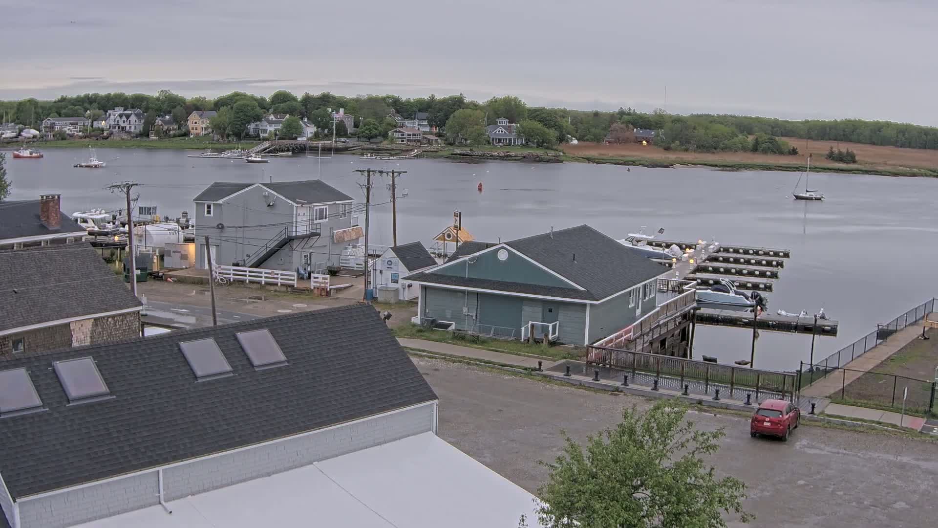 A harbor scene on an overcast day shows several buildings near a calm body of water with boats moored at a dock.