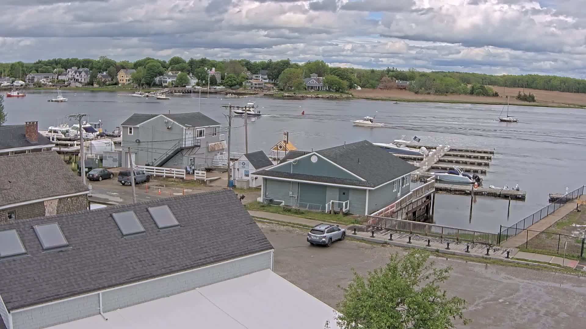 A waterfront scene under a cloudy sky shows a marina with boats docked, several buildings, cars in a parking lot, and a river with more boats and a shoreline with houses and trees in the distance.
