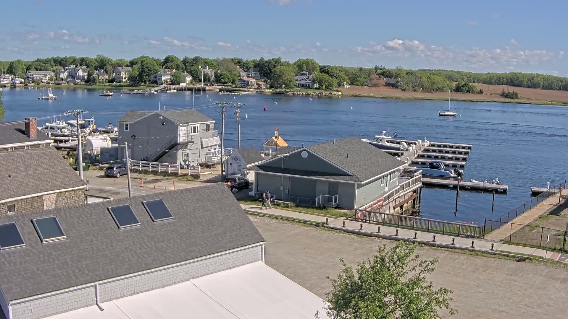 A waterfront scene on a sunny day shows several buildings, boats docked at a marina, and a calm river with a few other boats sailing.