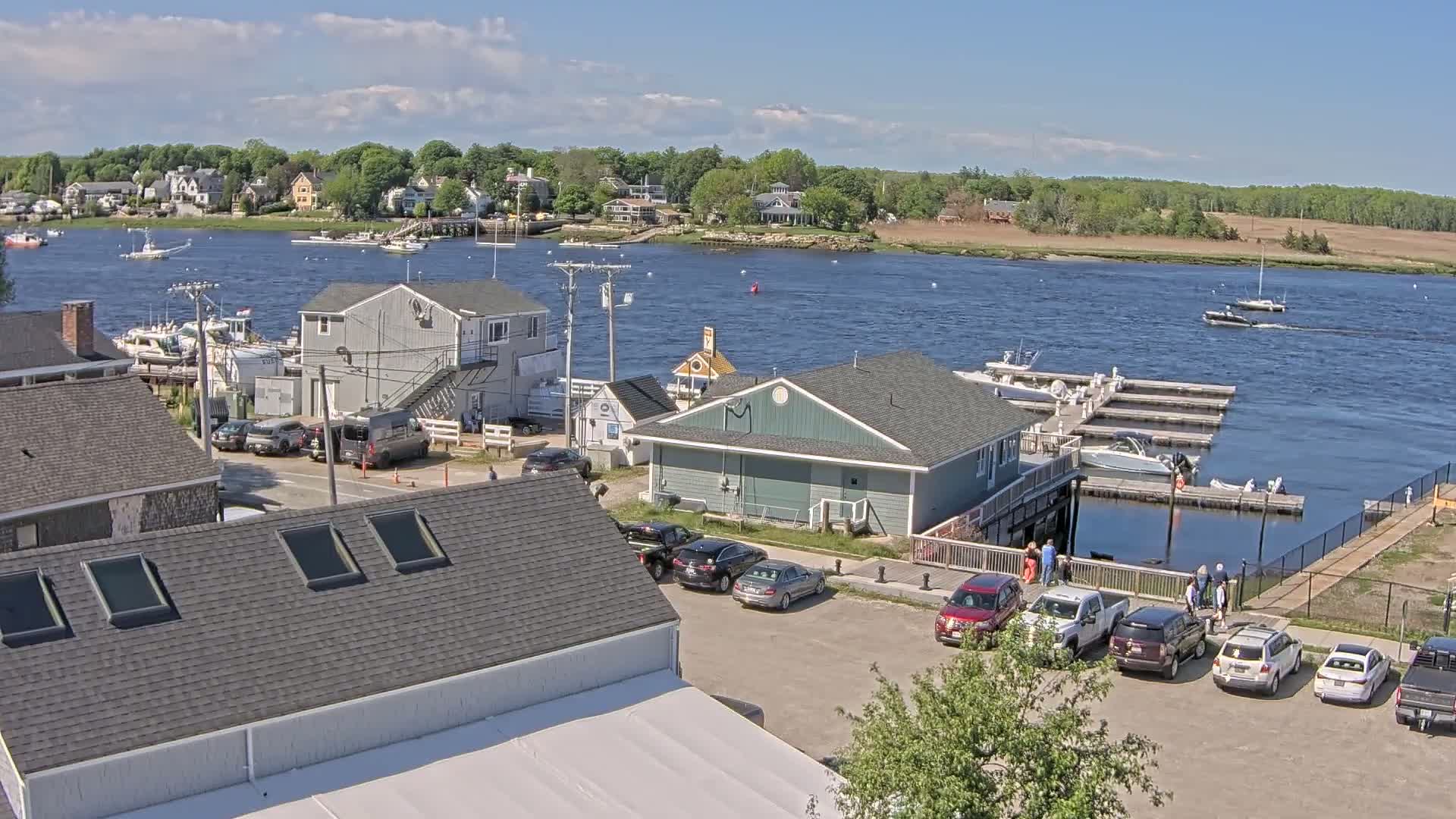 A waterfront scene on a partly sunny day shows a marina with boats docked, several cars parked in a lot, and houses along a river with trees and other houses visible in the background.