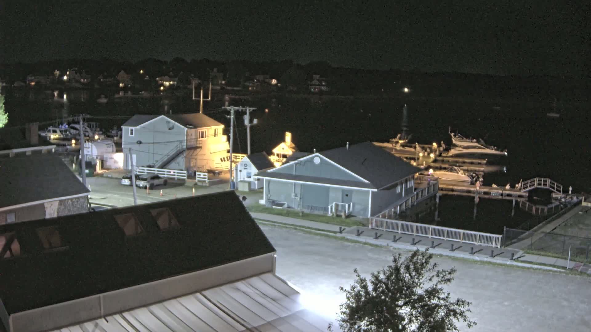 A nighttime aerial view shows a waterfront area with several buildings, boats docked at piers, and a small tree near a street.