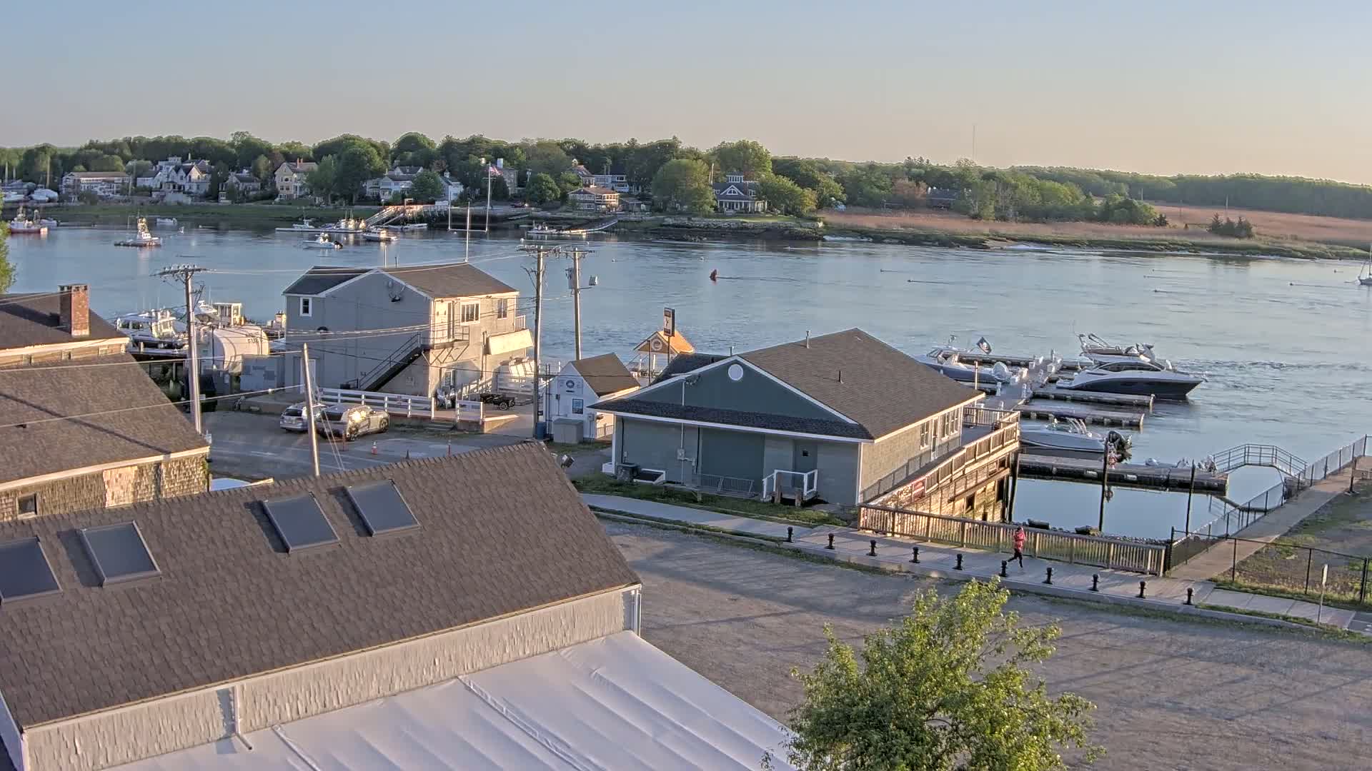 A waterfront town at sunset, with several boats docked at a marina, houses lining the riverbank, and a person walking along a paved path.