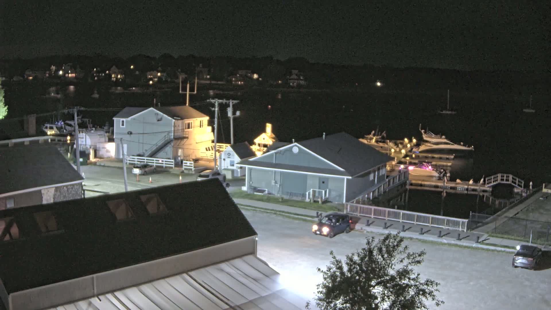 A nighttime view of a marina with several boats docked, lit buildings, and a few cars parked on a street near the water.