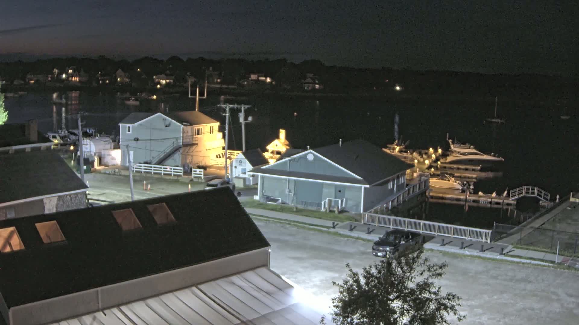 At night, a waterfront area shows several buildings, docked boats, and a few cars in a parking lot under clear skies.