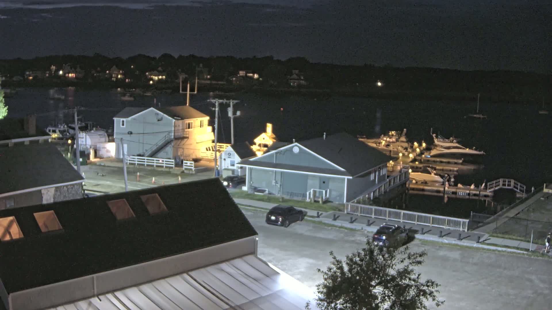 A nighttime view of a marina with docked boats, several buildings, and cars parked along a street, under a partly cloudy sky.