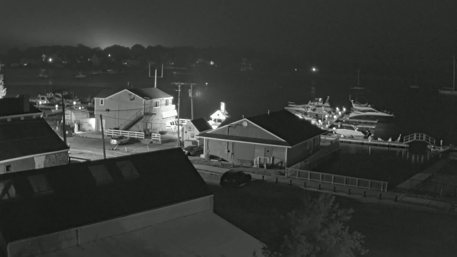 A nighttime, foggy view of a small harbor with boats docked, buildings, and cars parked along a street.