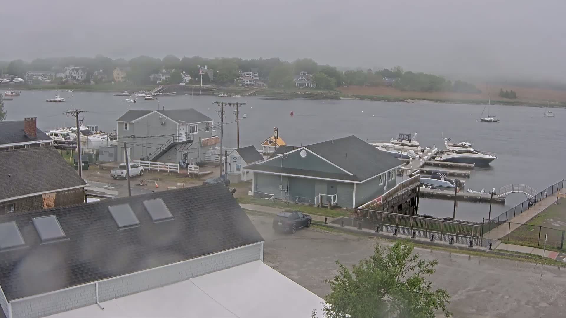 A waterfront town scene on a foggy, overcast day shows several buildings, a marina with boats docked, and a calm body of water with a few boats in the distance.