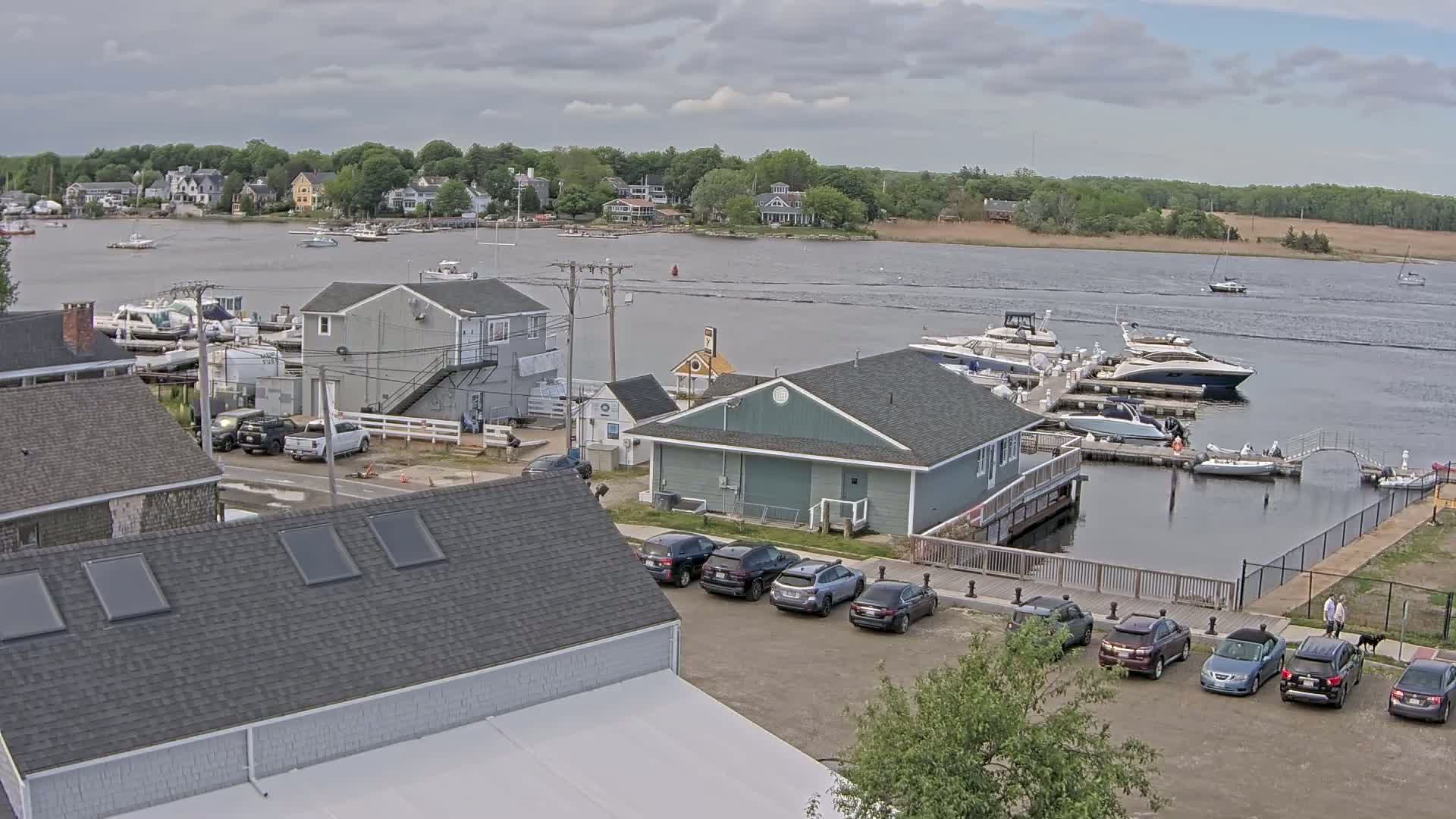 A waterfront scene on a partly cloudy day shows a marina with several boats docked, buildings, and cars parked in a lot.