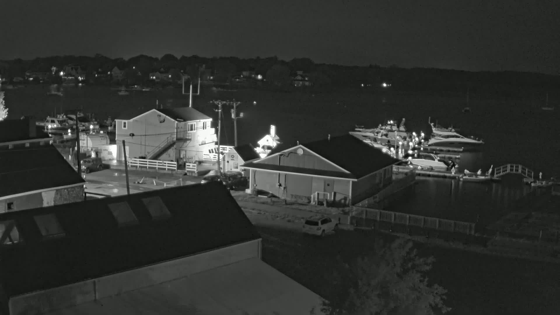 A nighttime, grayscale view of a waterfront area showing several buildings, docked boats, and vehicles parked along a roadway.
