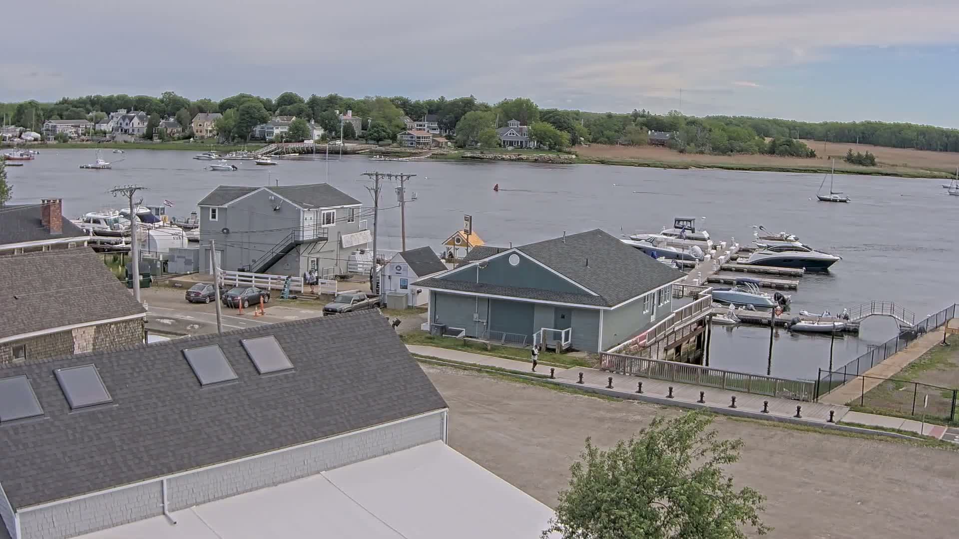 A waterfront scene on an overcast day shows a marina with several boats docked, a few buildings, and houses in the background across a calm river.