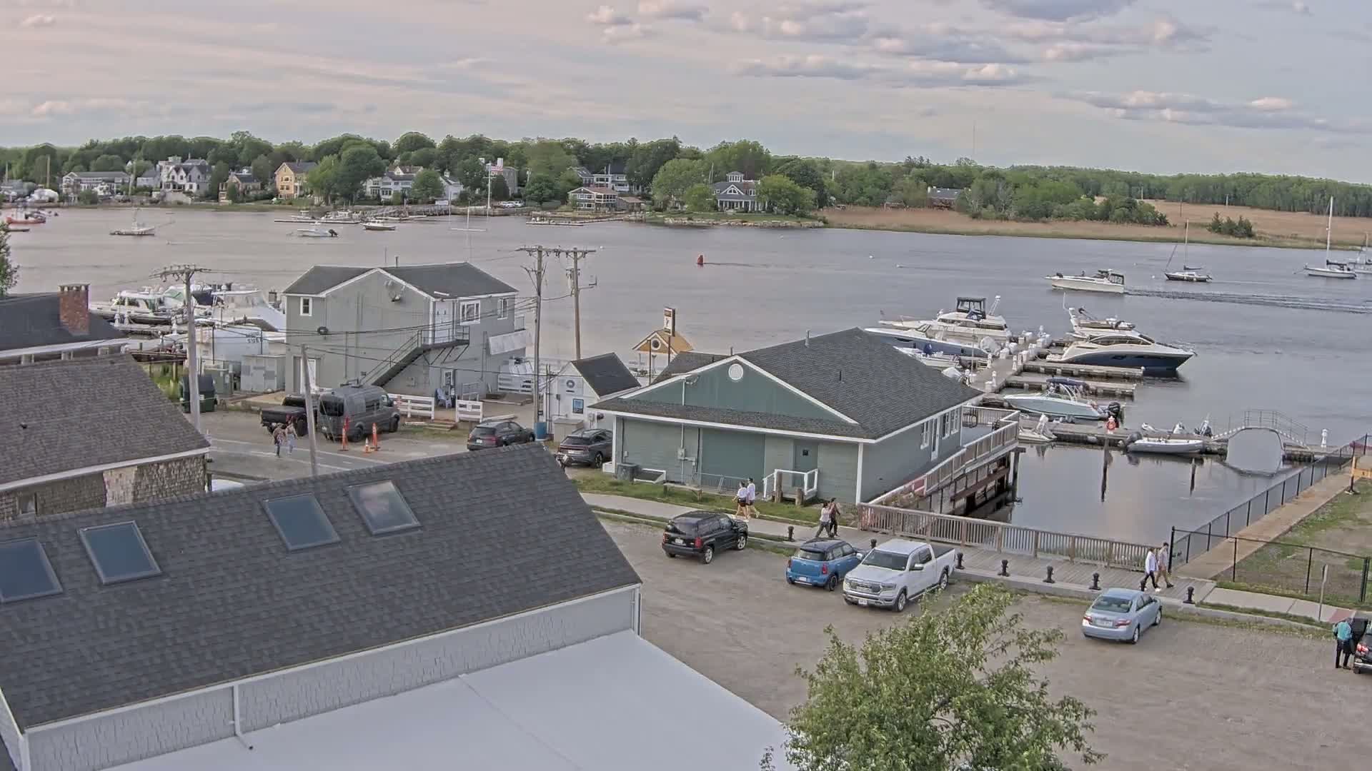 A marina with several boats docked, surrounded by buildings and a parking lot, under a partly cloudy sky.