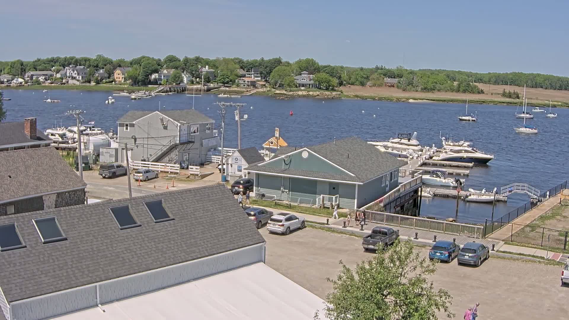 A marina with numerous boats docked, surrounded by buildings and parked cars, on a sunny day.