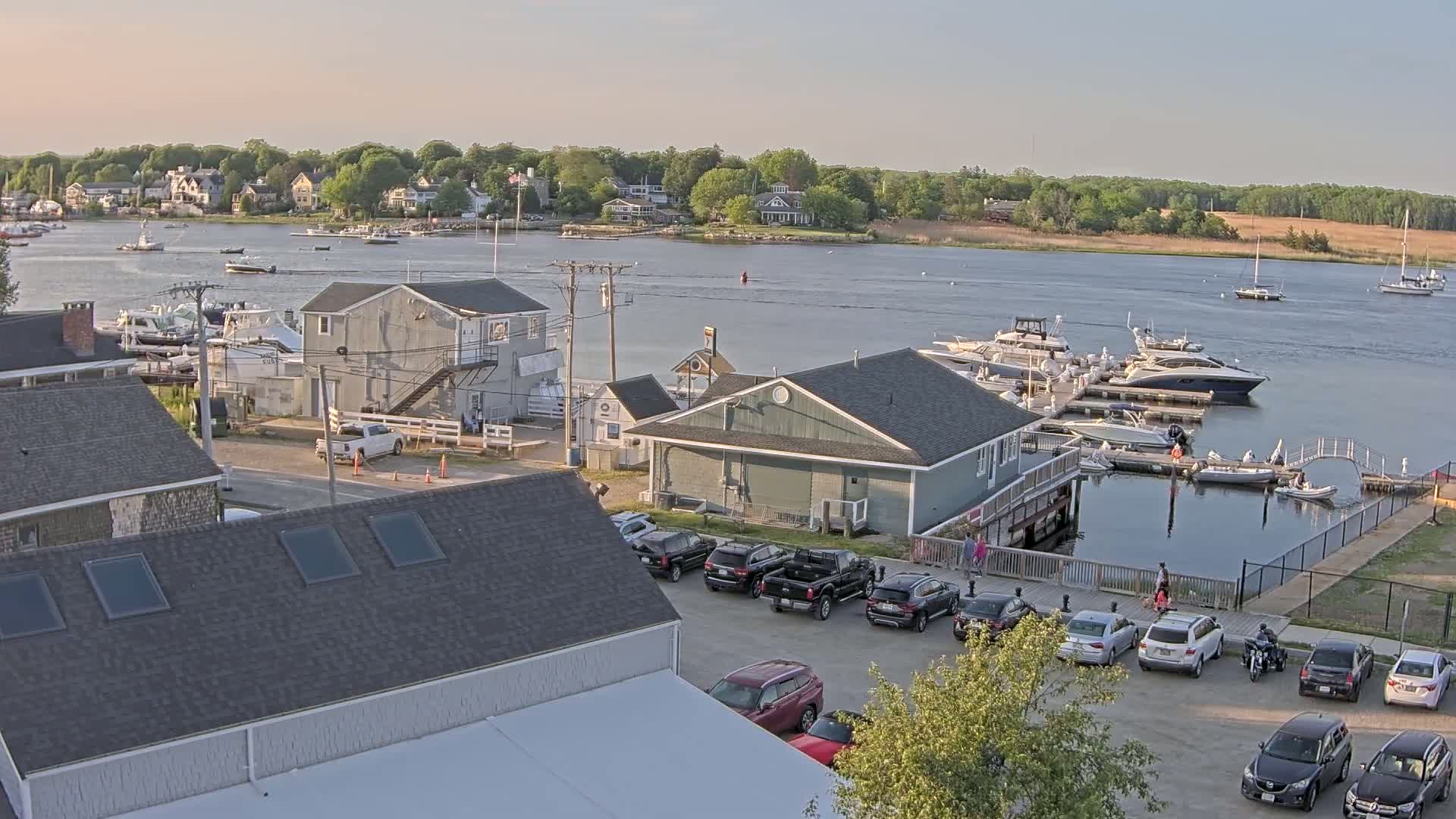 A marina with numerous boats docked, surrounded by buildings and cars, under a partly cloudy sky.