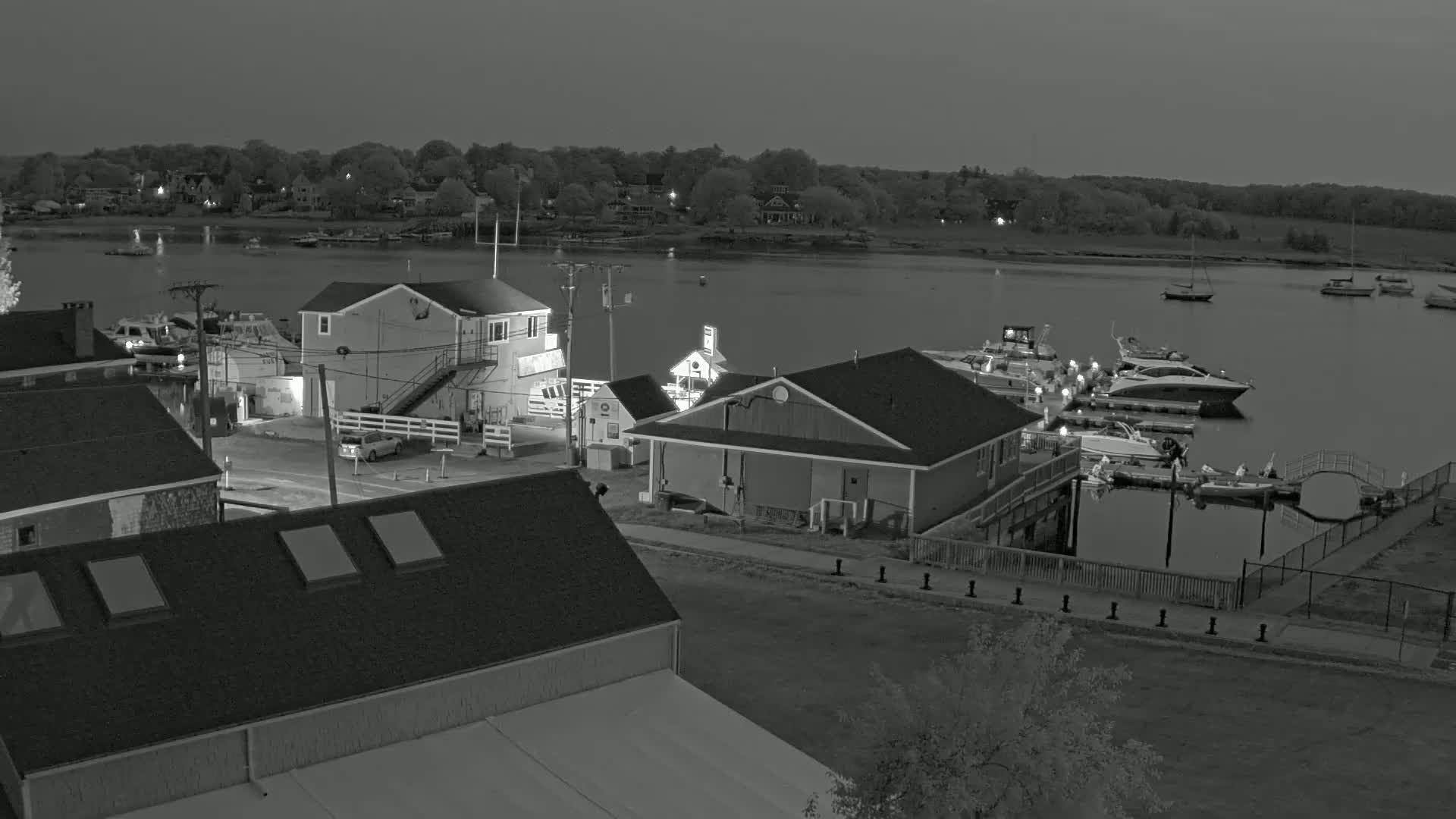 A nighttime, grayscale view of a small marina with several boats docked, adjacent buildings, and a calm body of water beyond.