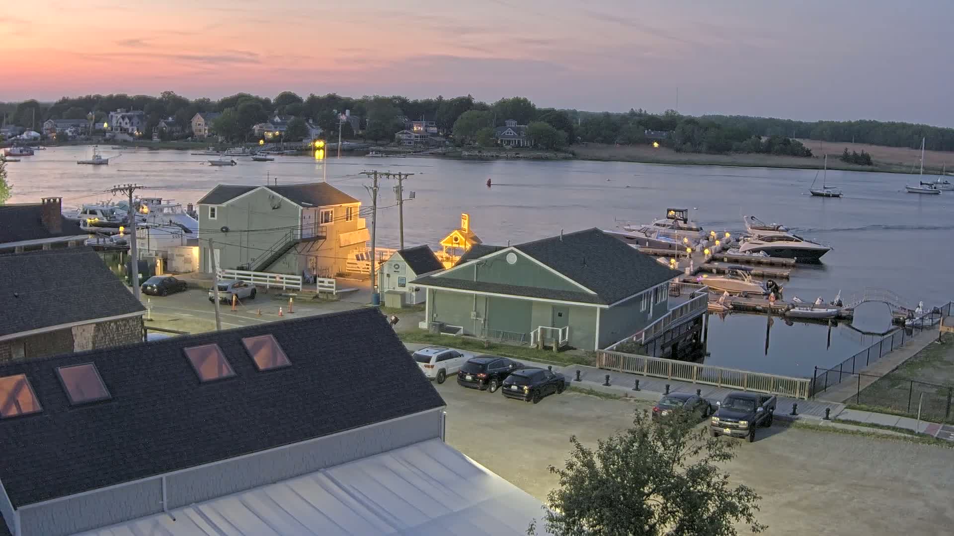 A waterfront scene at sunset shows several buildings, cars in a parking lot, and numerous boats docked at a marina.