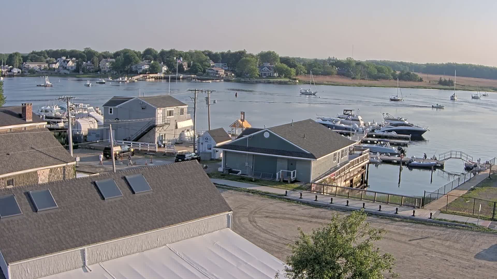 A calm, sunny day reveals a waterfront scene with docked boats, houses, and a small town across a river.