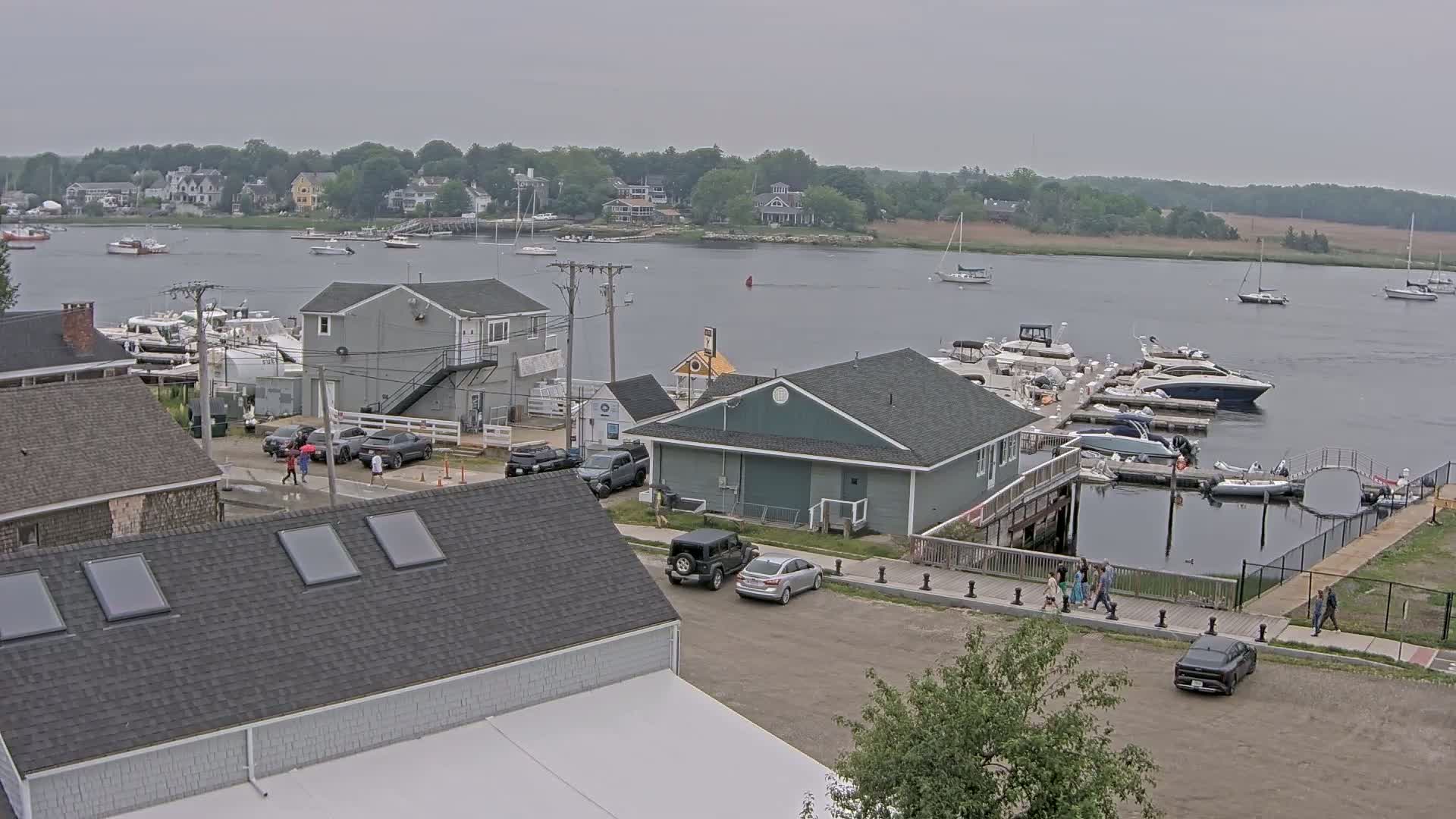 An overcast day reveals a marina with numerous boats docked, surrounded by buildings and a few people walking near a parking lot.