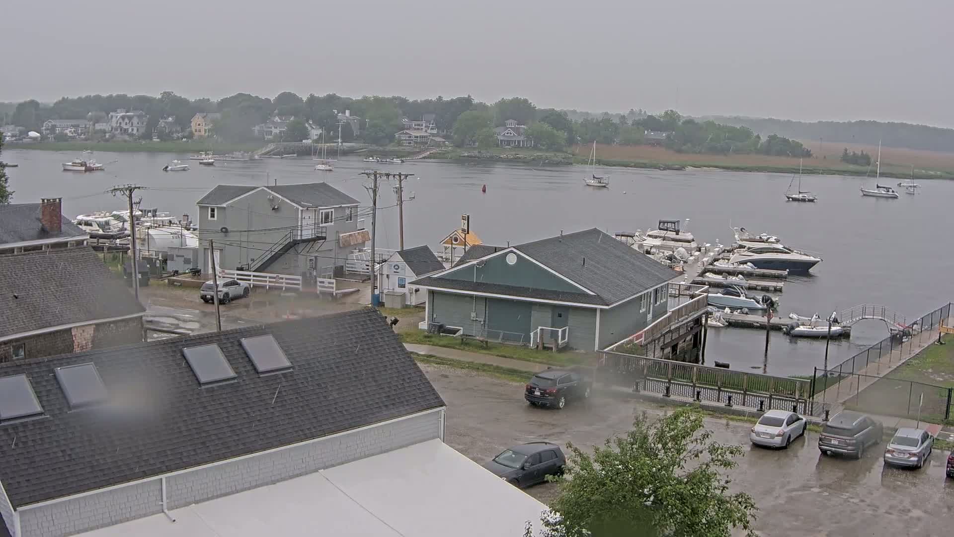 A rain-soaked waterfront scene shows a marina filled with boats, several buildings, and cars parked in a lot, with houses visible across a calm body of water in the background.