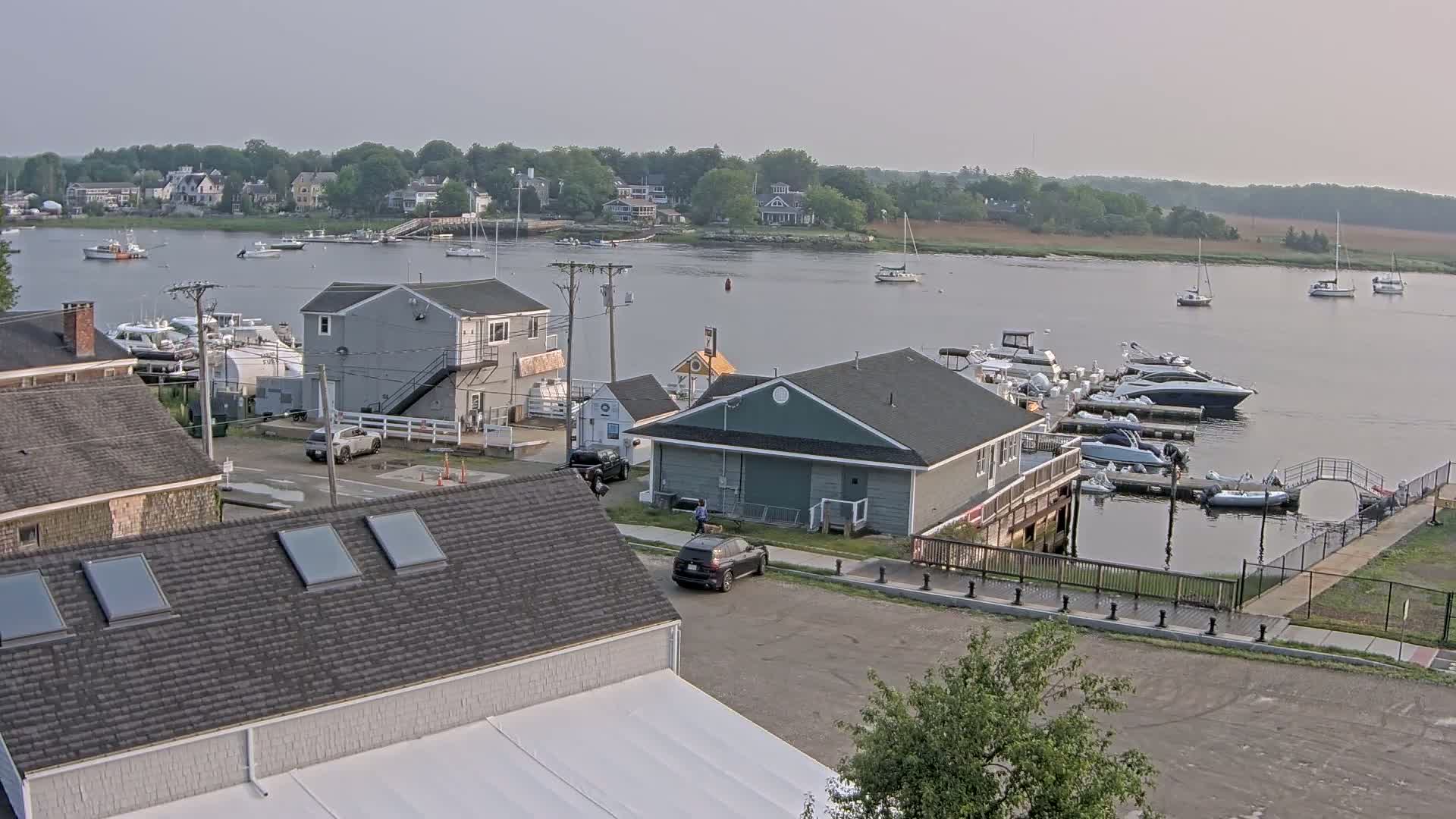 A waterfront scene on a calm, hazy day shows a marina with numerous boats, several buildings, and a few cars parked nearby.