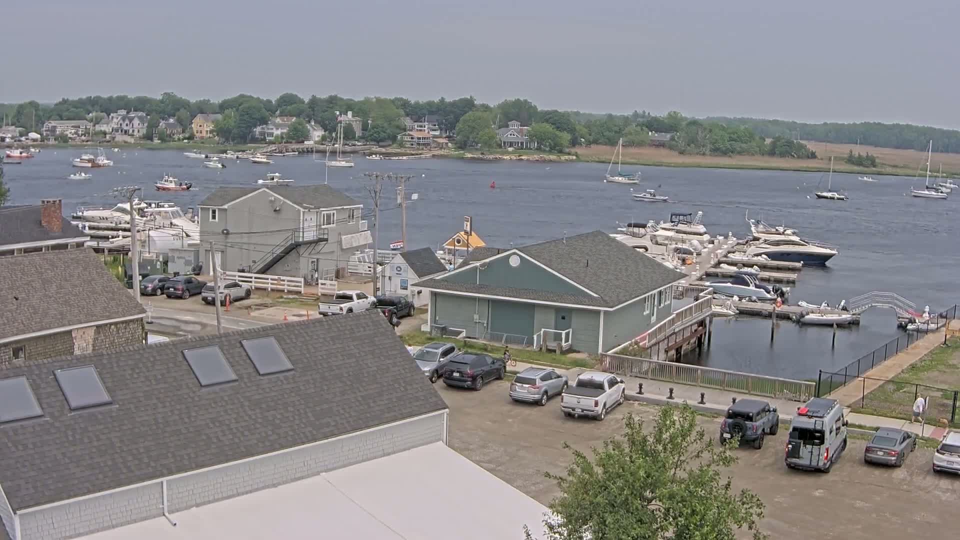 A waterfront view on an overcast day shows a marina filled with boats, adjacent buildings, and a parking lot with several cars and a van.