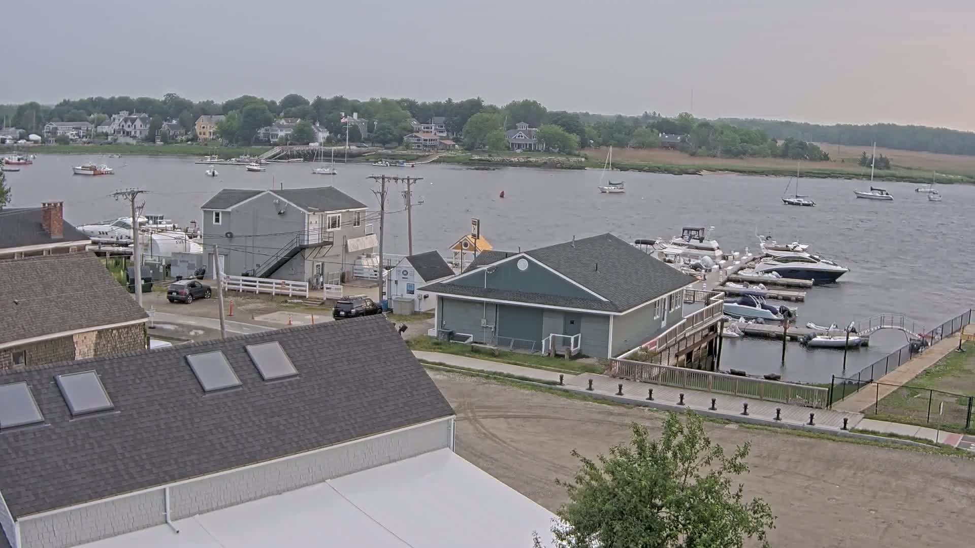 A marina with several boats docked, alongside waterfront buildings and houses, under an overcast sky.