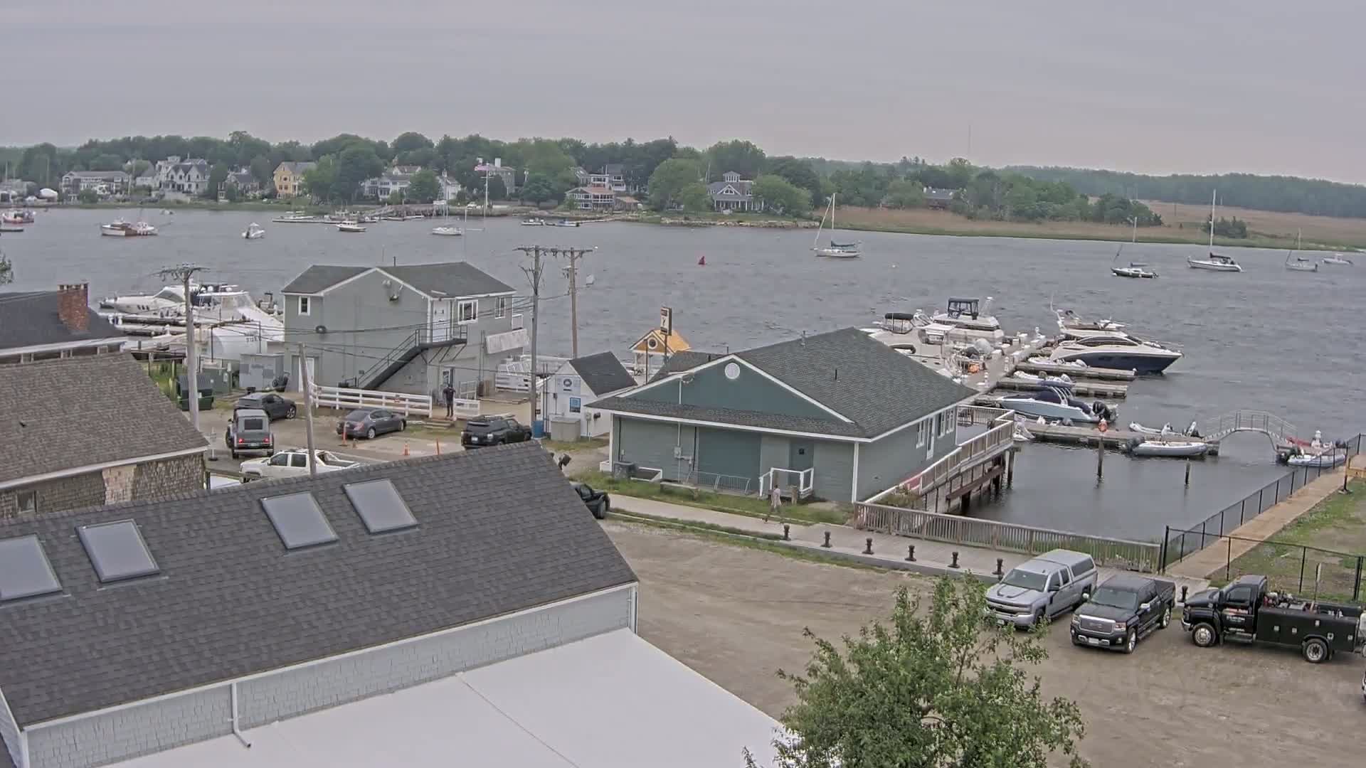 A marina with numerous boats docked, along with several buildings and parked vehicles, under a cloudy sky.