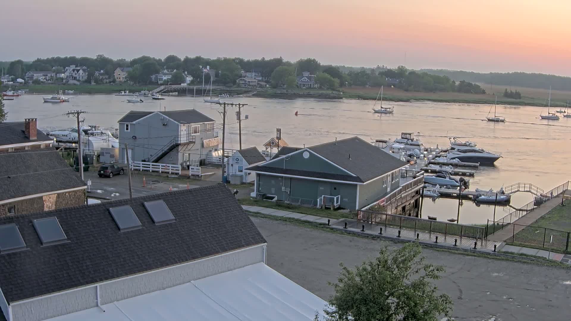 A waterfront town at sunrise shows a calm river with numerous boats and yachts docked at a marina, alongside buildings and a parking lot.