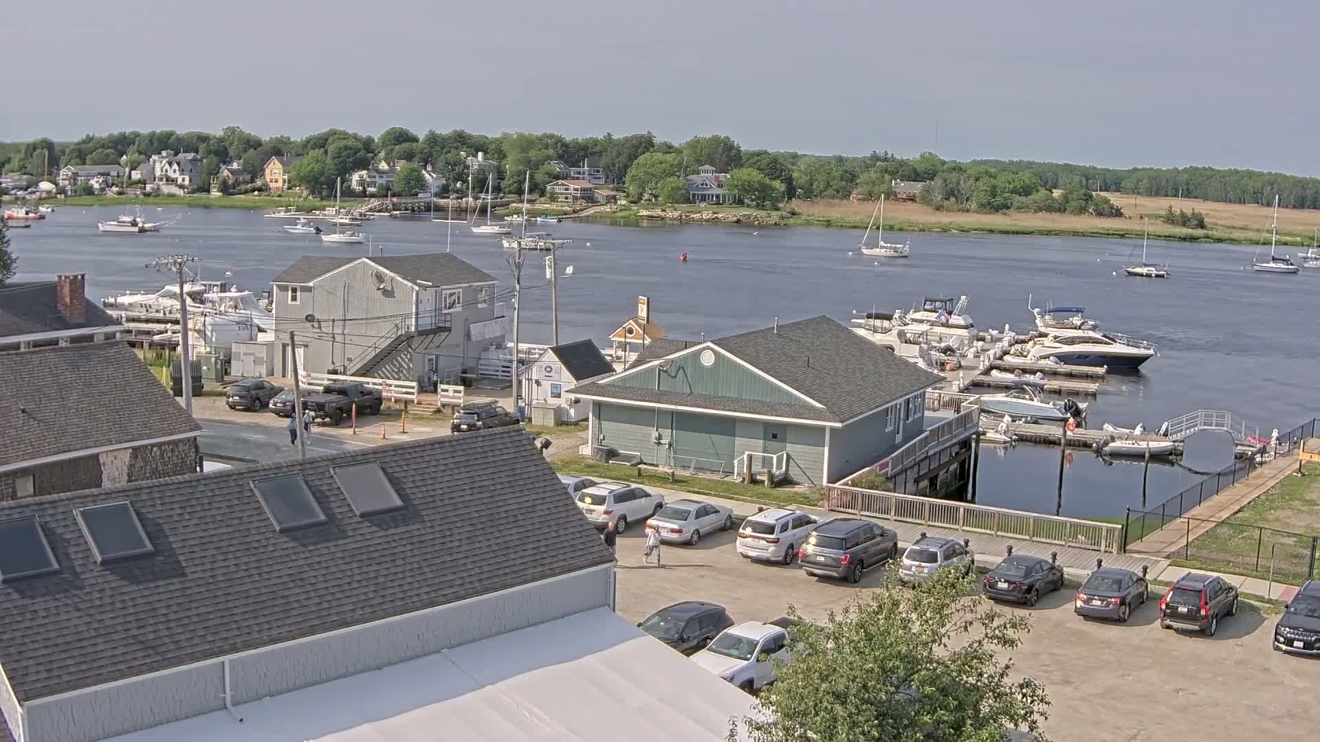 A sunny day overlooking a calm river with numerous boats docked and moored, alongside a parking lot filled with cars near waterfront buildings.