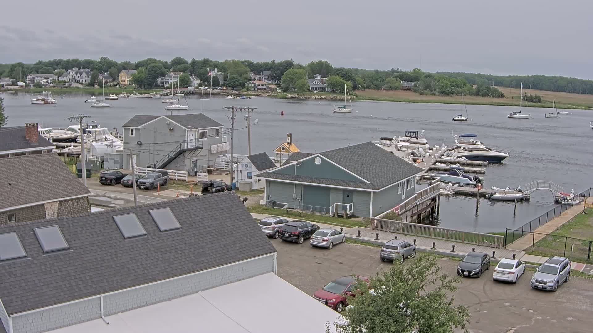 A waterfront scene on an overcast day shows a marina with several boats docked, a parking lot with cars, and several buildings, including houses and commercial structures.