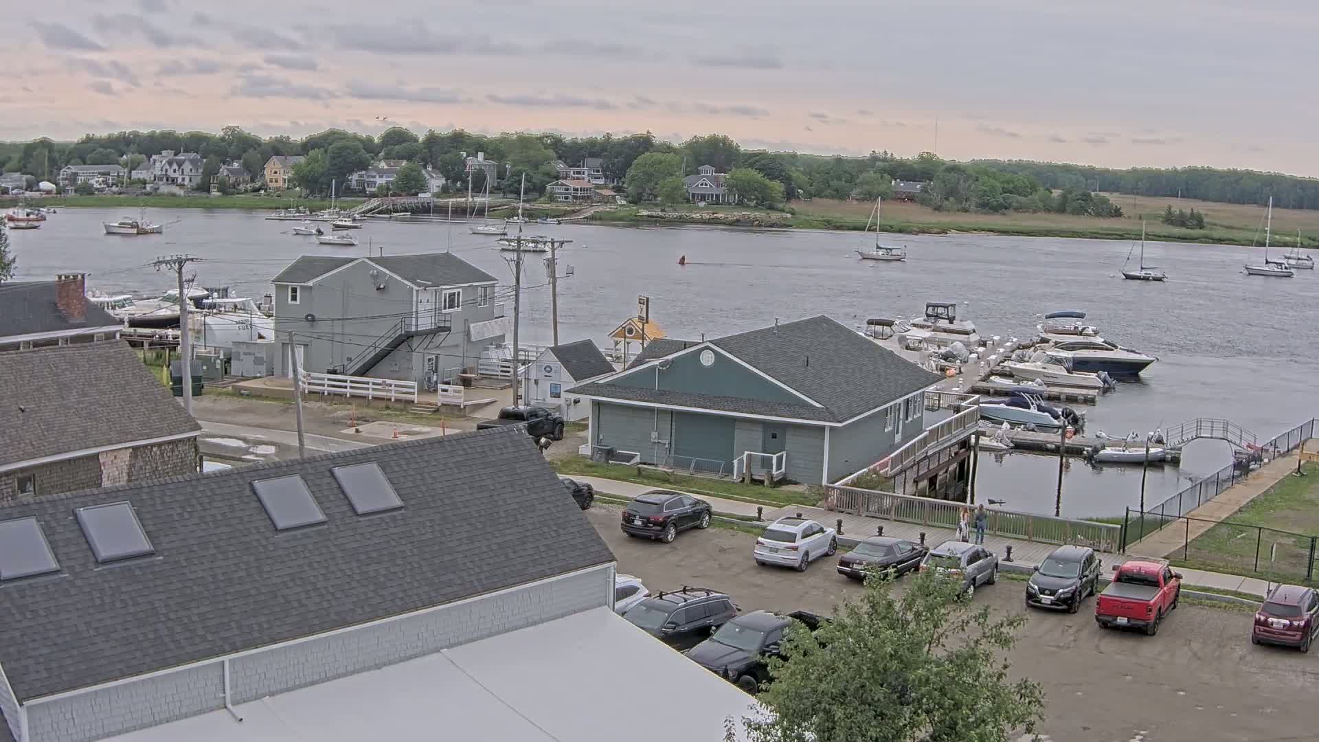 A waterfront scene on an overcast day shows a marina with several boats docked, a parking lot with cars, and several buildings, including houses and commercial structures.