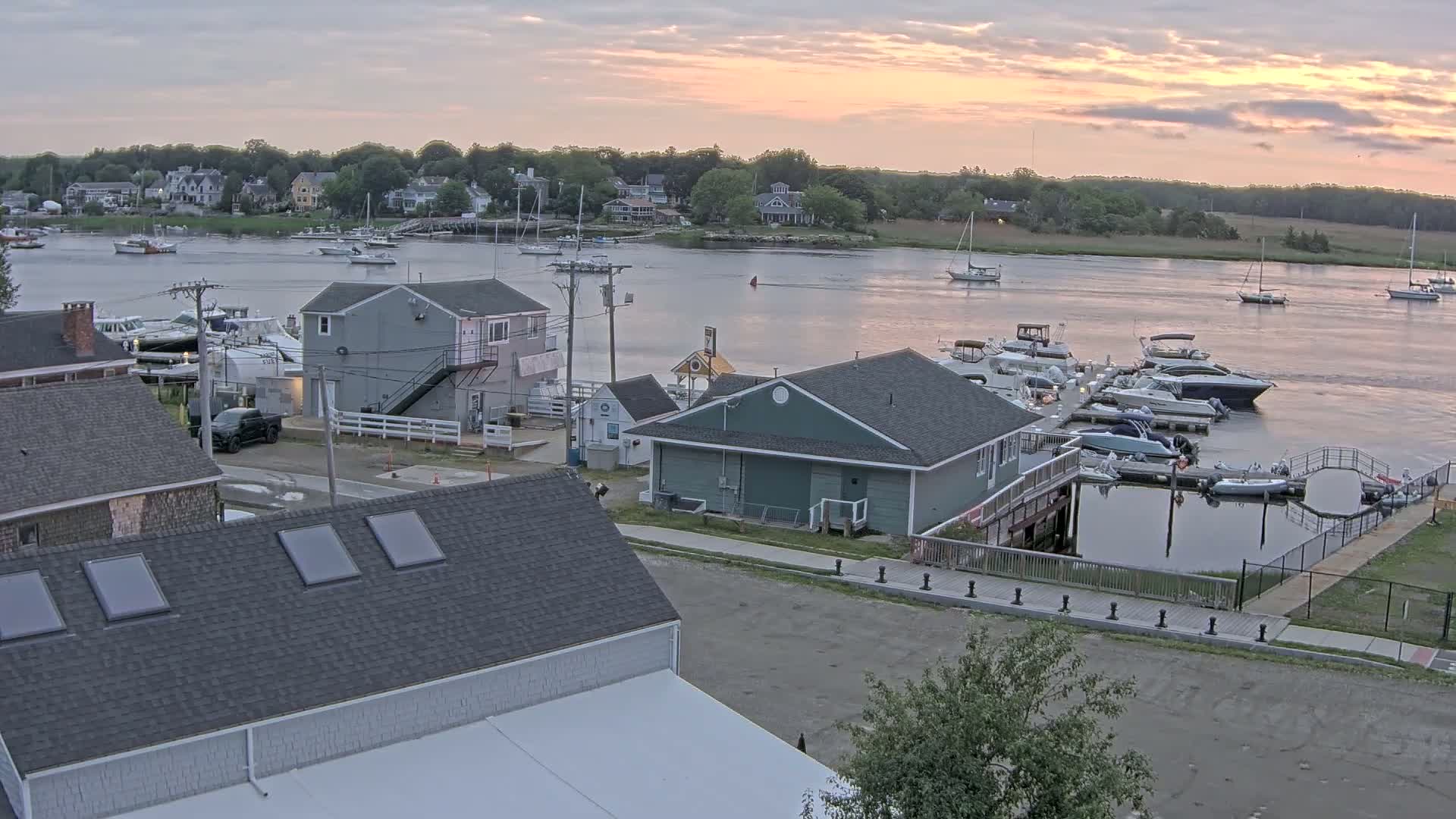 A waterfront scene at sunrise shows several buildings, docked boats, and calm water with some boats sailing in the distance.