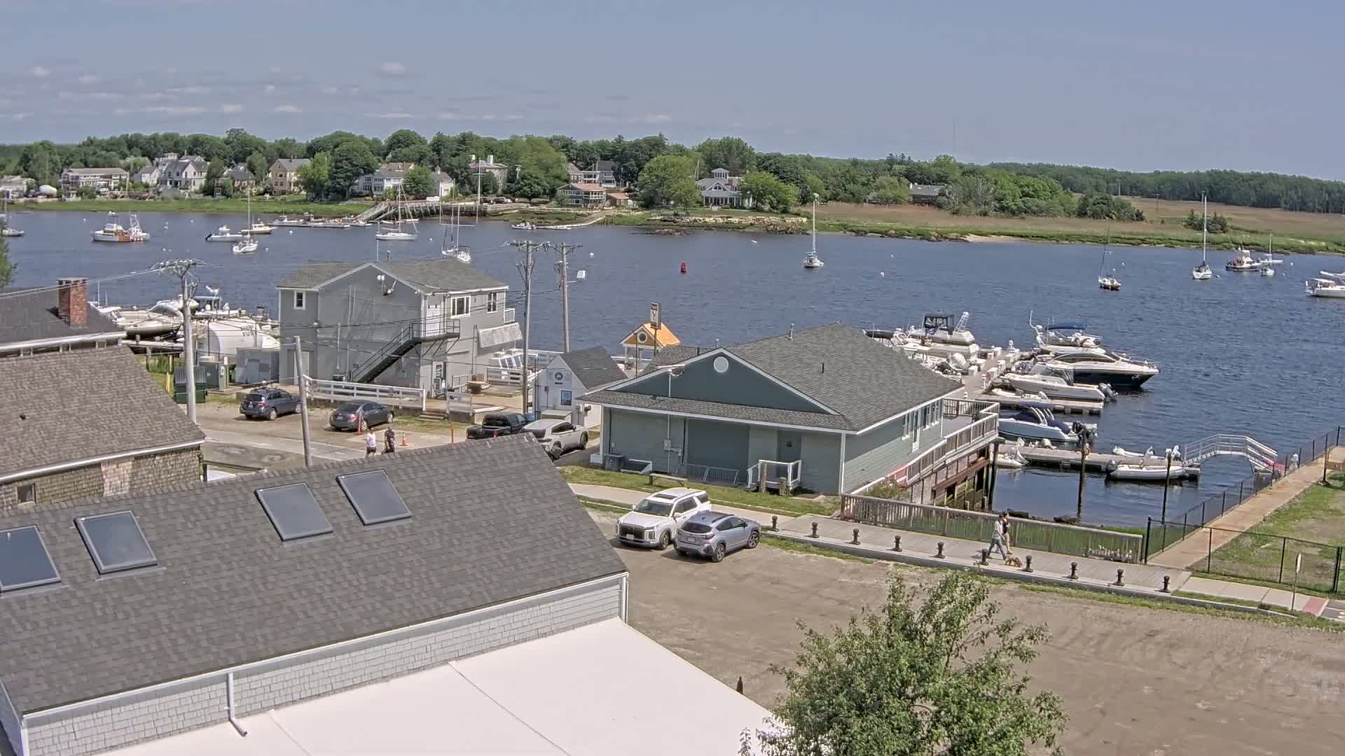 A waterfront scene on a sunny day shows a marina filled with boats, houses lining the shore, and people walking near a parking lot.