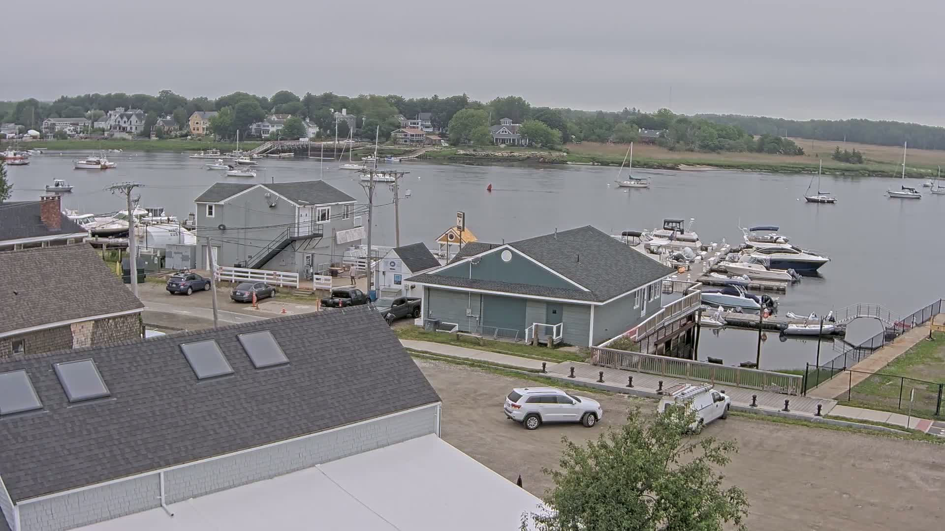A waterfront scene on an overcast day shows a marina filled with boats, several houses and buildings, and a few cars in a parking lot.