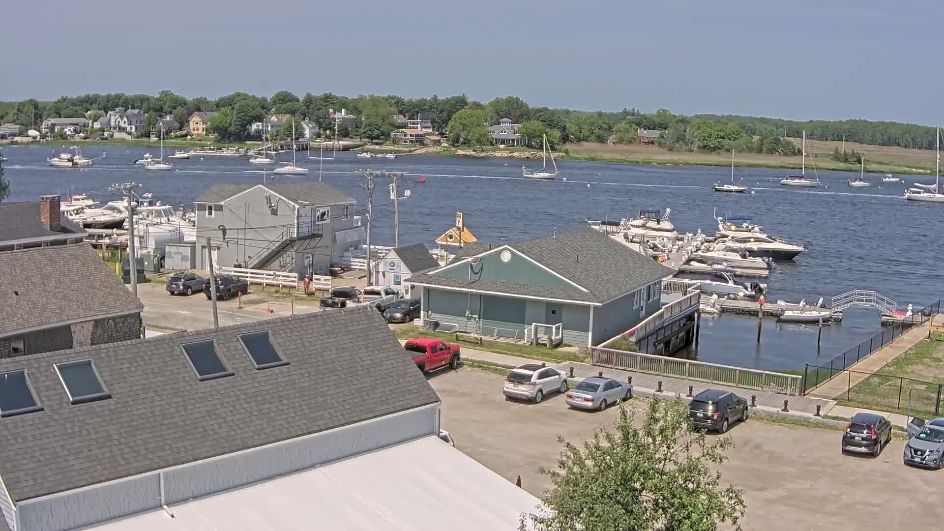 A sunny day at a marina shows numerous boats docked and moored in the water, with several cars parked in a lot next to buildings on the shore.