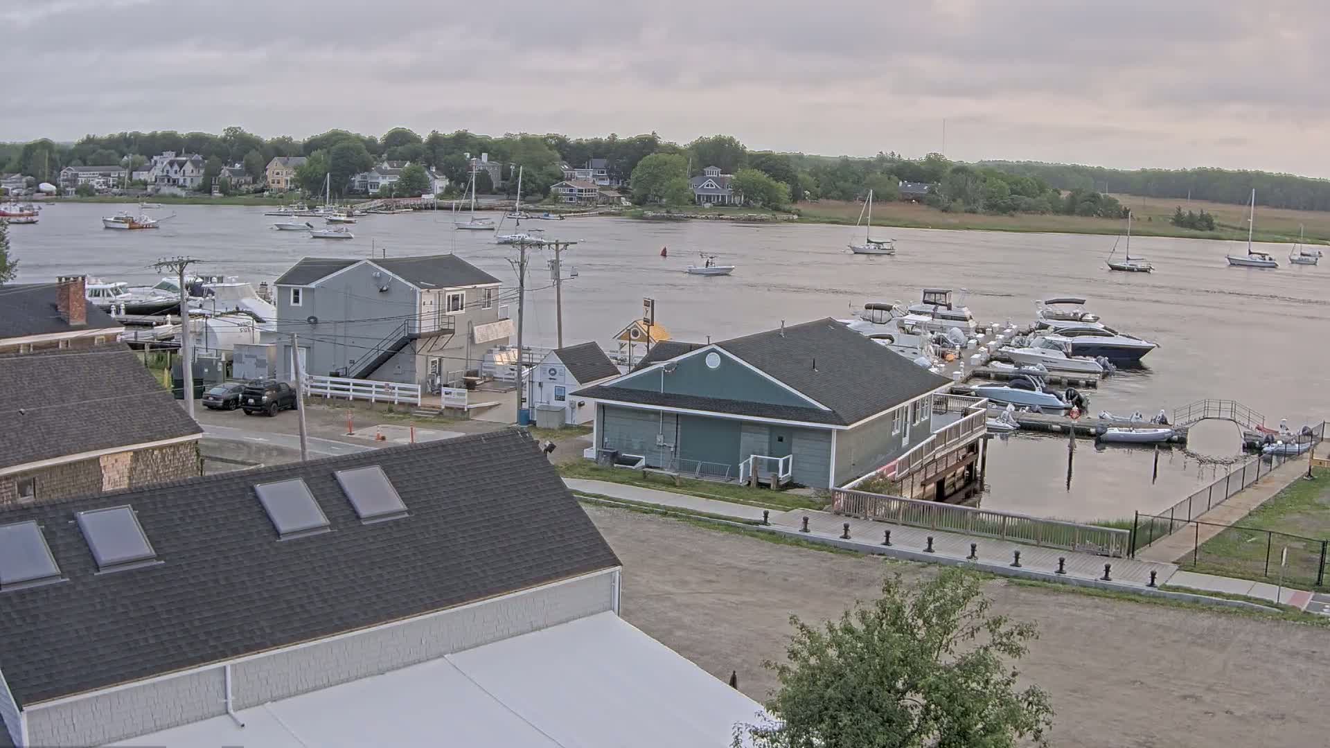 A calm, overcast day shows a marina filled with boats, alongside waterfront buildings and houses across a bay.
