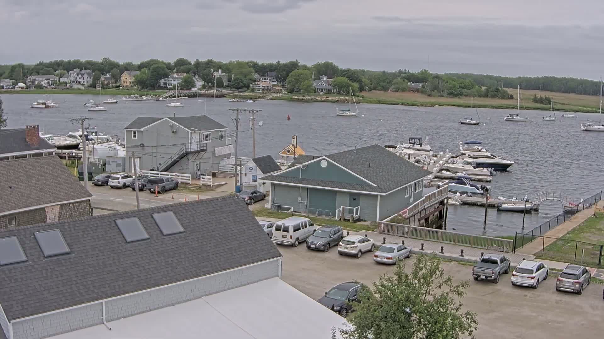 A waterfront scene on an overcast day shows a marina with numerous boats docked, a parking lot with several cars and vans, and a collection of houses and buildings along the shore.